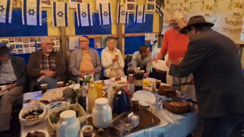Deelnemers zitten aan een gedekte tafel in de versierde Soeka, met Isra&euml;lische vlaggetjes en foto's aan de wand