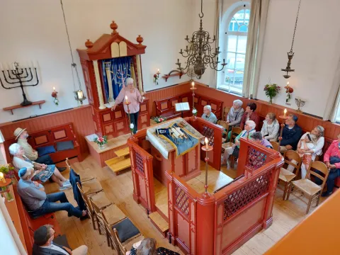 Bovenaanzicht van de synagogezaal: een rondleidster vertelt bij de Thora-ark aan bezoekers in de banken; op de bima ligt een Thorarol