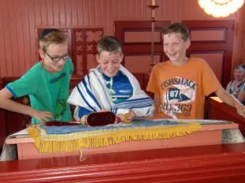 Drie lachende jongens bij de bima; de middelste draagt een gebedsmantel en leest uit een Thorarol
