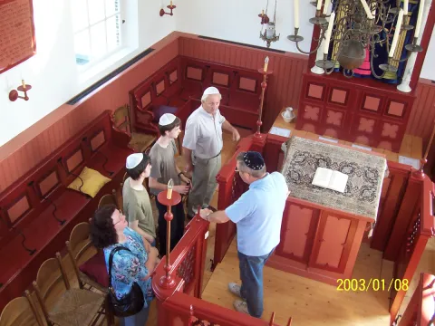 Een gids met keppeltje geeft een klein gezelschap uitleg naast de bima in de synagogezaal