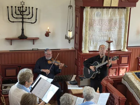 Bovenaanzicht van koorleden met zangmappen in de Dijksynagoge, met viool en gitaar voor de Heilige Ark