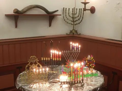 Brandende chanoekia's van verschillende stijlen op een tafel in de hoek van de synagoge, met een sjofar op een plank erboven