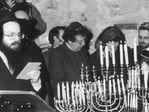 Zwart-witfoto van een rabbijn die voorleest, met bezoekers achter brandende chanoekia's, december 1997