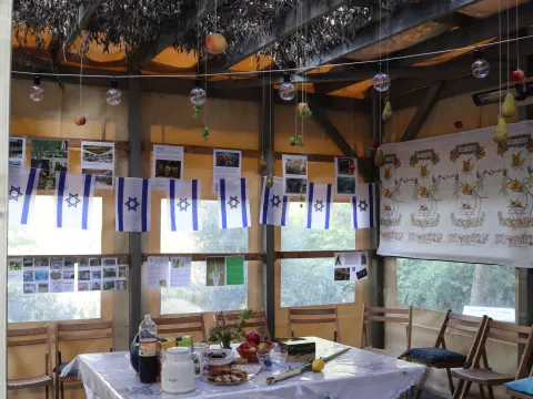 Interieur van de Soeka met een gedekte tafel op een kleed, houten stoelen, Isra&euml;lische vlaggetjes en een loofdak met hangende vruchten