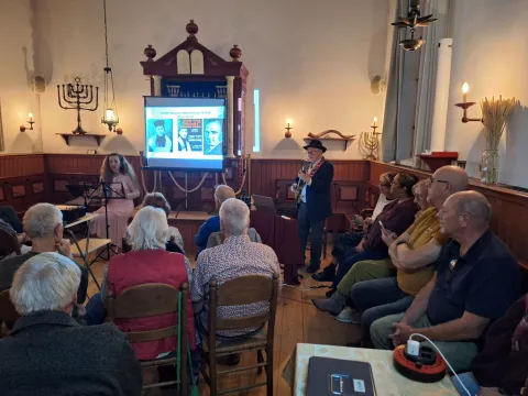 Flip de Liema met gitaar en zangeres Orsaya treden op in de synagoge voor een volle zaal, met beelden op een scherm voor de Heilige Ark