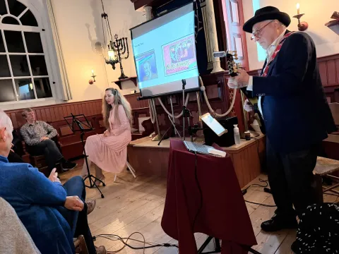 Flip de Liema met hoed speelt gitaar naast een scherm met beelden, terwijl zangeres Orsaya in een roze jurk toekijkt
