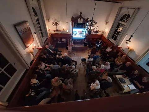 De sfeervol verlichte synagoge van bovenaf gezien, met bezoekers in een kring rond de optredenden en het scherm voor de Heilige Ark