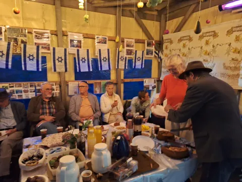 Deelnemers zitten aan een gedekte tafel in de versierde Soeka, met Isra&euml;lische vlaggetjes en foto's aan de wand